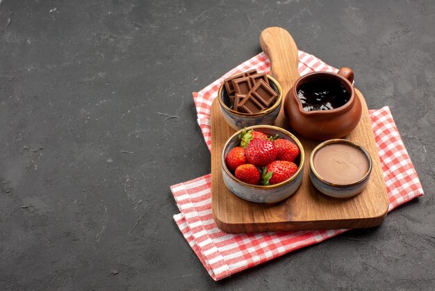 Side close-up view bowls on board bowls of chocolate cream and strawberries on the wooden cutting board on the pink-white checkered tablecloth on the right side of the dark table