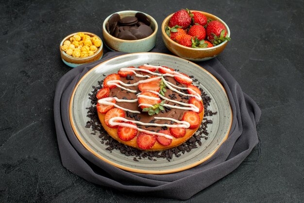 Side close-up view cake on tablecloth pie with chocolate and strawberry pieces on grey tablecloth next to bowls of hazelnut strawberry and chocolate and cake on black table