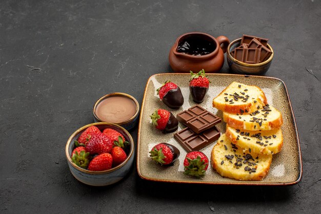 Side close-up view cake with strawberries tasty cake with chocolate-covered strawberries and chocolate cream strawberry and chocolate in bowls on the dark background