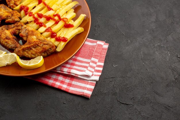 Side close-up view chicken french fries on checkered tablecloth orange plate of appetizing french fries chicken wings ketchup and lemon on the left side of the dark table