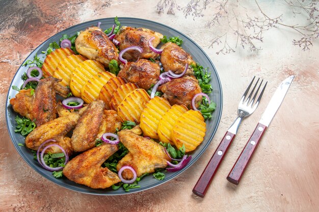 Вид сбоку крупным планом куриная тарелка с куриными крылышками, картофель, зелень, лук, вилка, нож