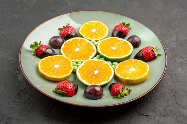 Vista ravvicinata laterale frutti ricoperti di cioccolato fragole ricoperte di cioccolato caramelle verdi e appetitosa arancia tritata al centro del tavolo nero