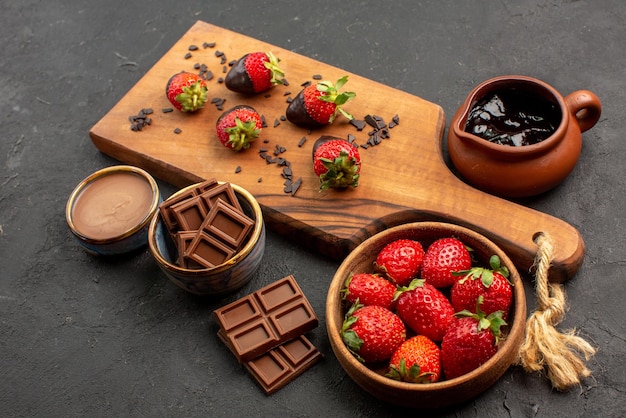 Side close-up view chocolate starwberries on board bars of chocolate chocolate cream strawberries in bowl next to chocolate-covered strawberries on the kitchen cutting board