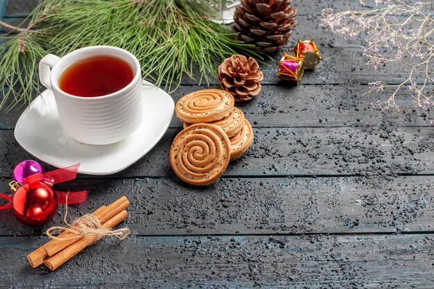 Biscotti di vista ravvicinata laterale biscotti di natale accanto all'albero di abete con coni e giocattoli di natale bastoncini di cannella una tazza di tè sul piattino sul tavolo di legno