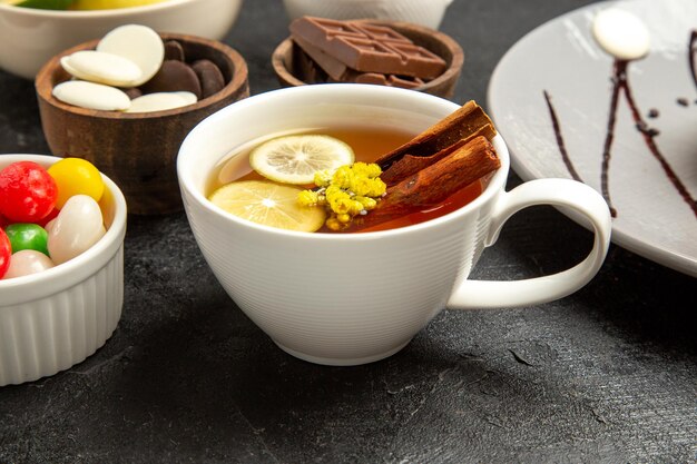 Side close-up view a cup of tea a cup of tea with lemon and cinnamon sticks next to the bowls of candies on the dark table