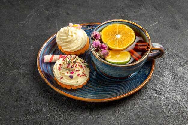 Side close-up view a cup of tea with lemon two cupcakes with cream and sweets next to the cup of herbal tea with lemon and cinnamon on the dark table