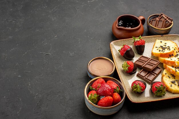 Side close-up view dessert cake with chocolate-covered strawberries and chocolate with bowls of chocolate cream and berries on the right side of the dark table