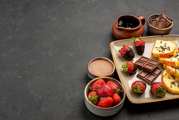 Side close-up view dessert cake with chocolate-covered strawberries and chocolate with bowls of chocolate cream and berries on the right side of the dark table