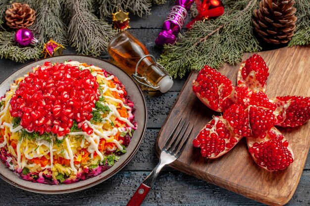 Side close-up view dish and spices dish with pomegranate potatoes next to the bottle of oil pilled pomegranate fork on the cutting board and tree branches with cones and Christmas tree toys