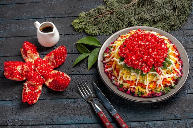 Free photo side close-up view dish with pomegranates dish next to the pilled pomegranate oil next to the knife and fork spruce branches on the dark table