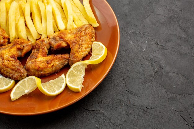 Side close-up view fastfood orange plate of an appetizing chicken wings french fries and lemon on the dark background