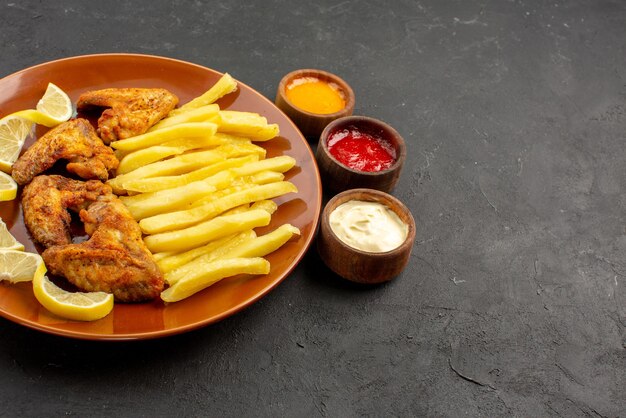 Side close-up view fastfood orange plate of chicken wings appetizing french fries and lemon and three bowls of different types of sauces on the dark table