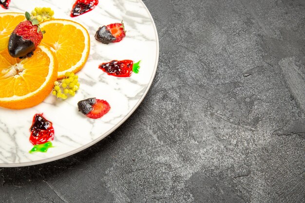 Side close-up view fruits on the plate chocolate-covered strawberries and sliced orange on the white plate on the dark table