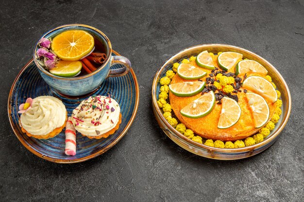 Side close-up view herbal tea blue cup of herbal tea with cinnamon sticks and lemon and two cupcakes with cream next to the plate of an appetizing cake with candies and limes on the black table