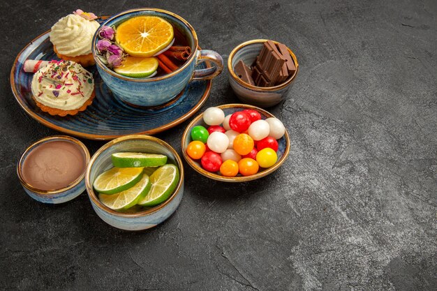 Side close-up view herbal tea bowls of chocolate cream slices of lime colorful candies next to the cup of tea with lemon and two cupcakes with cream on the black table