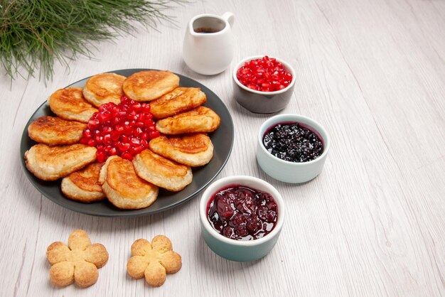 Боковой вид крупным планом блины, блины и гранат в тарелке рядом с мисками с ягодами, печеньем, соусом и елкой на столе