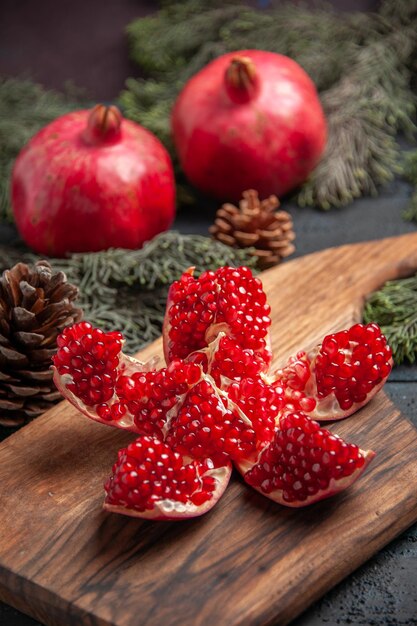 Side close-up view pomegranate red pomegranate on wooden board next to spruce branches with cones and two red pomegranates on grey background