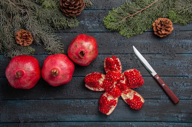 Side close-up view pomegranates on table pilled pomegranate next to three red pomegranates knife and spruce branches with cones