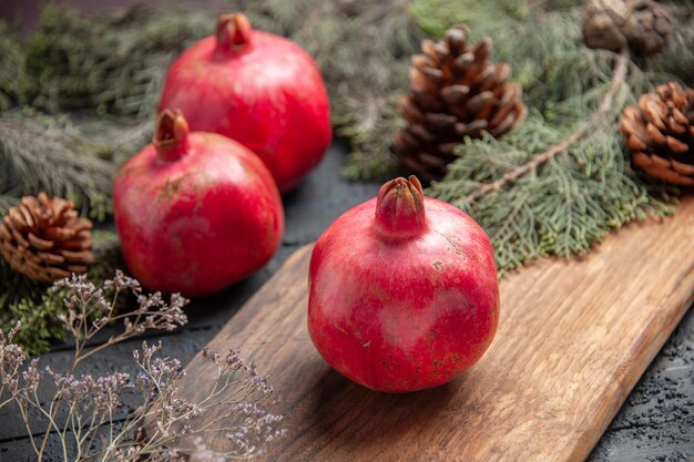 Side close-up view ripe pomegranates and branches red pomegranate on cutting board next to two pomegranates and spruce branches with cones on grey table