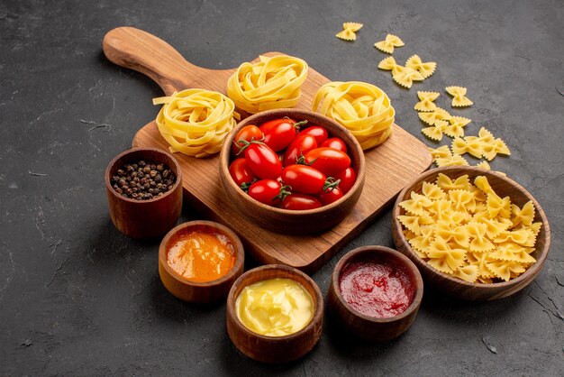 Side close-up view sauces pasta pasta and colorful sauces black pepper next to the bowl of tomatoes and pasta on the kitchen board on the table