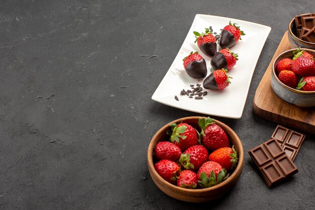 Side close-up view strawberries on board chocolate cream and strawberries on the board next to the plate of chocolate-covered strawberries and bowl of strawberries