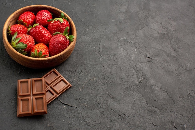 Free photo side close-up view strawberries in bowl appetizing bars of chocolate next to the strawberries in bowl on the left side of the dark table