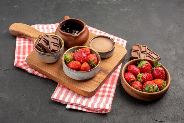 Side close-up view strawberries on tablecloth kitchen board with bowls of strawberries and chocolate on it and plate of strawberries and chocolate in the center of the table