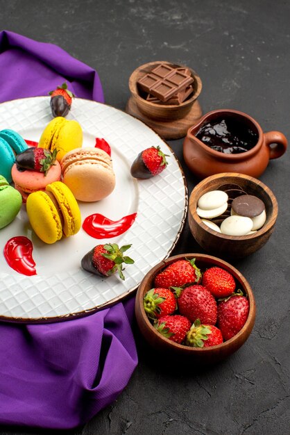 Side close-up view sweets French macaroons on the purple tablecloth and four bowls of sweets on the dark wall