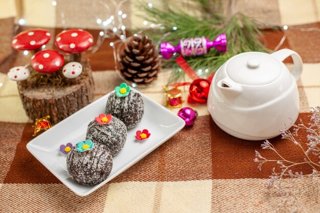 Side close-up view sweets with chocolate white teapot a cup of tea on a saucer next to the plate of chocolate sweets and tree branches with Christmas tree toys on the checkered tablecloth