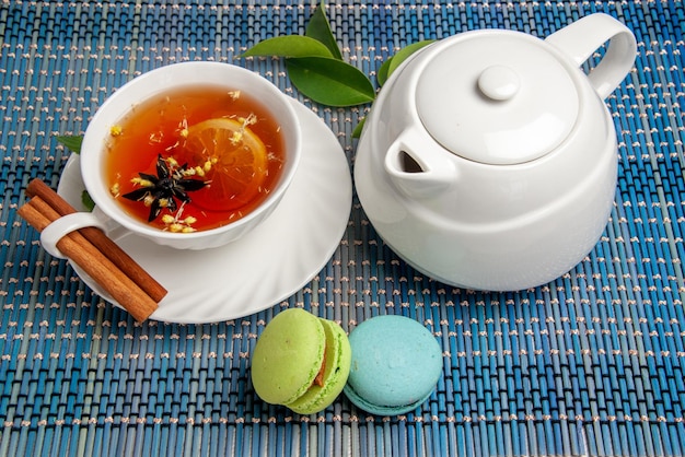 Free photo side close-up view teapot macaroons a cup of tea cinnamon sticks next to the teapot and french macaroons on the blue-white tablecloth