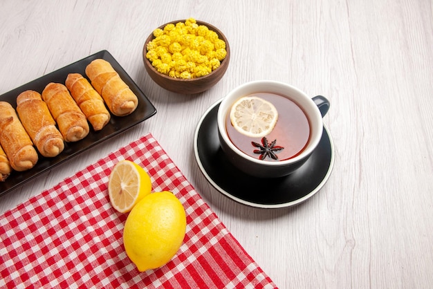 Крупным планом вид сбоку трубчатое тесто с лимоном на клетчатой скатерти темная тарелка с тестом рядом с миской желтых конфет и чашкой чая на белом столе