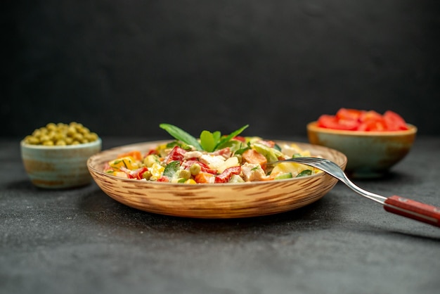 Side close view of bowl of vegetable salad with fork and bowls of vegetables on side on dark grey table on dark grey background