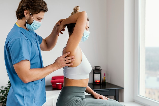 Foto gratuita lato del terapista osteopatico maschio con mascherina medica che controlla l'articolazione della spalla del paziente femminile