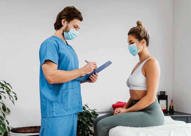 Side of male osteopathic therapist with medical mask and female patient in the office signing clipboard