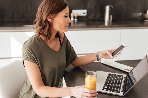 Side vew of casual woman using smartphone, tablet computer and drinking juice by the table on kitchen