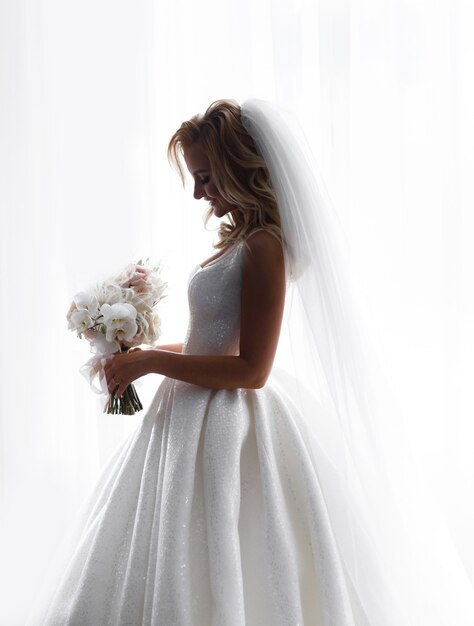 花を考慮した、結婚式の輝きのドレスとベールを身に着けている愛らしいフィアンセの側面図