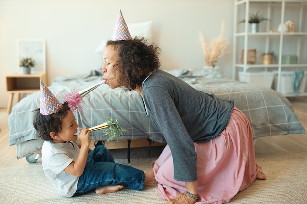 Free photo side view of adorable happy little boy sitting on floor with his young mother wearing cone hat, blowing whistles