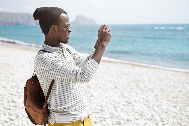 Free Photo | Side view of african american young man with backpack, in hat  and striped shirt taking photos of seaside standing on beach alone