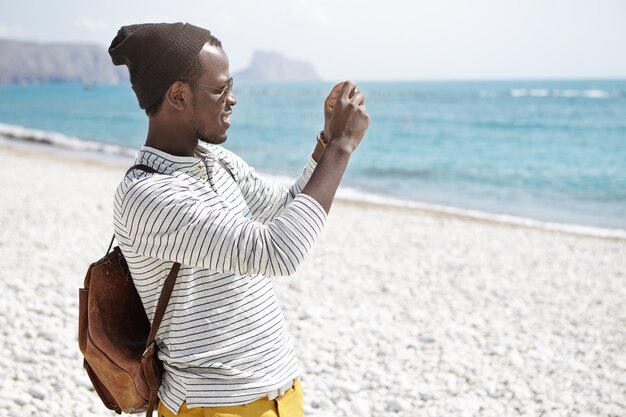 Side view of African American young man with backpack, in hat and striped shirt taking photos of seaside standing on beach alone