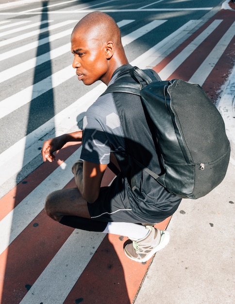 Foto gratuita vista laterale di un giovane uomo in buona salute africano con il suo zaino che si accovaccia sulla strada in città