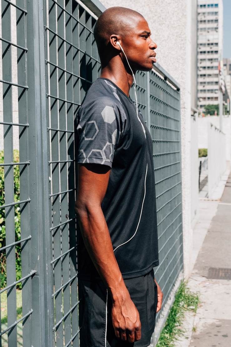 Free Photo side view of a african young male athlete listening music on earphones standing near the gate