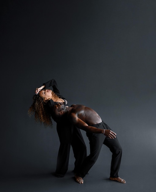 Side view of afro american man topless man and curly haires woman in suit bending bodies back while rehearsing of dance movement in isolated black studio