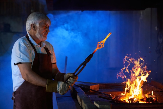 Side view of aged caucasian man in safety apron and gloves heating metal in burning fire at forge Experienced blacksmith using forceps while working with steel Manufacture concept