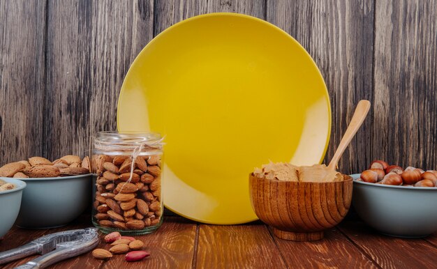 Side view of almond in a glass jar and a bowl with peanut butter in a bowl and yellow ceramic plate at wooden background