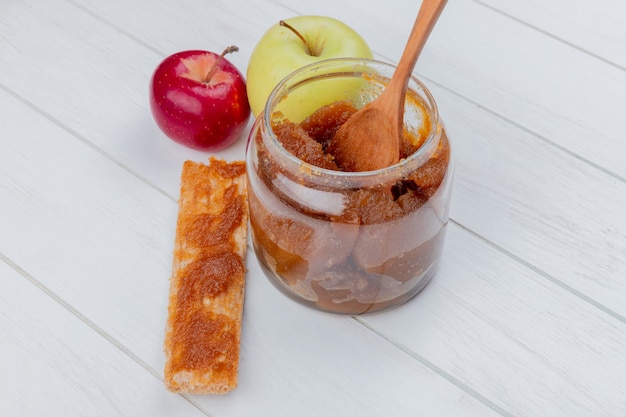 Side view of apple jam in glass jar and smeared on cookie with apples and wooden spoon on wooden surface