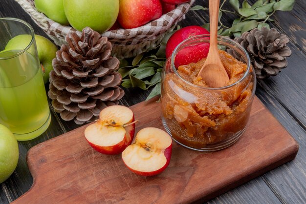 Side view of apple jam in glass jar with wooden spoon and half cut apple on cutting board with apple juice basket of apples pinecone and leaves on wooden surface