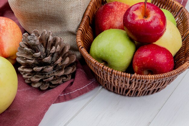 Side view of apples in basket with pinecone and apples on bordo cloth and wooden surface
