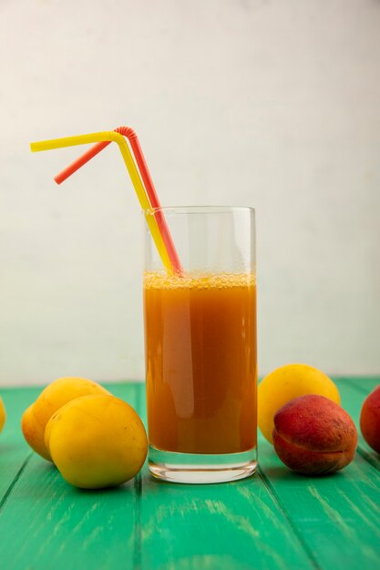 Вид сбоку абрикосового сока с питьевыми трубками в стекле и абрикосами на зеленой поверхности и белом фоне