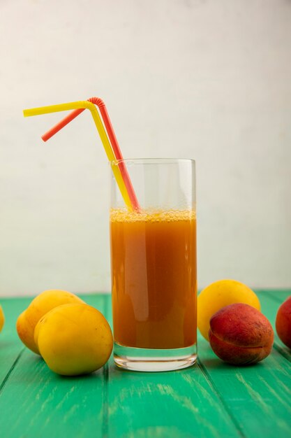 Side view of apricot juice with drinking tubes in glass and apricots around on green surface and white background