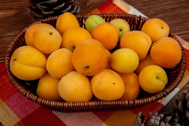 Side view of apricots in basket on cloth with pinecones on wooden background